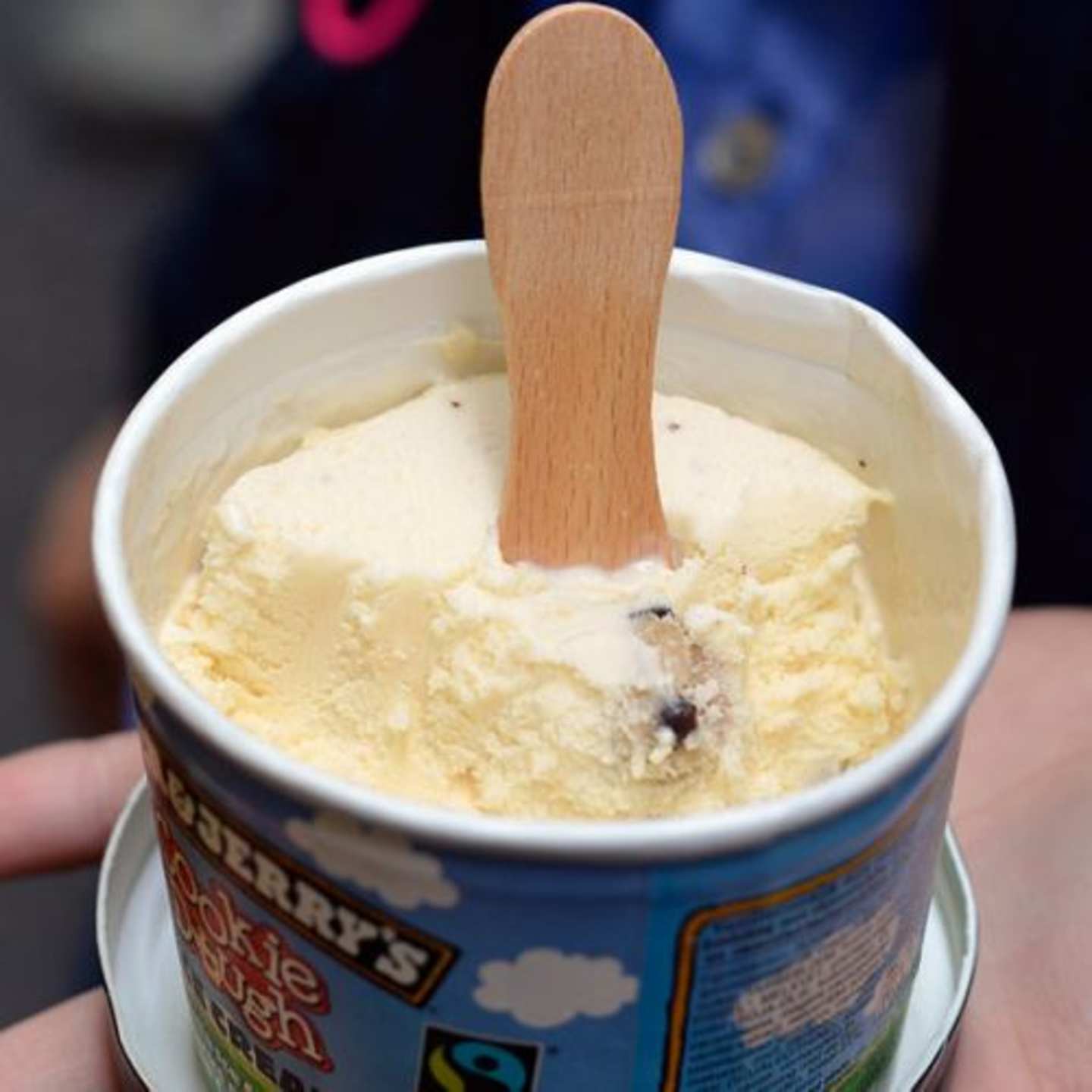 A white container holding a creamy, light-colored ice cream with a wooden spoon inserted into it, set against a blurred background.