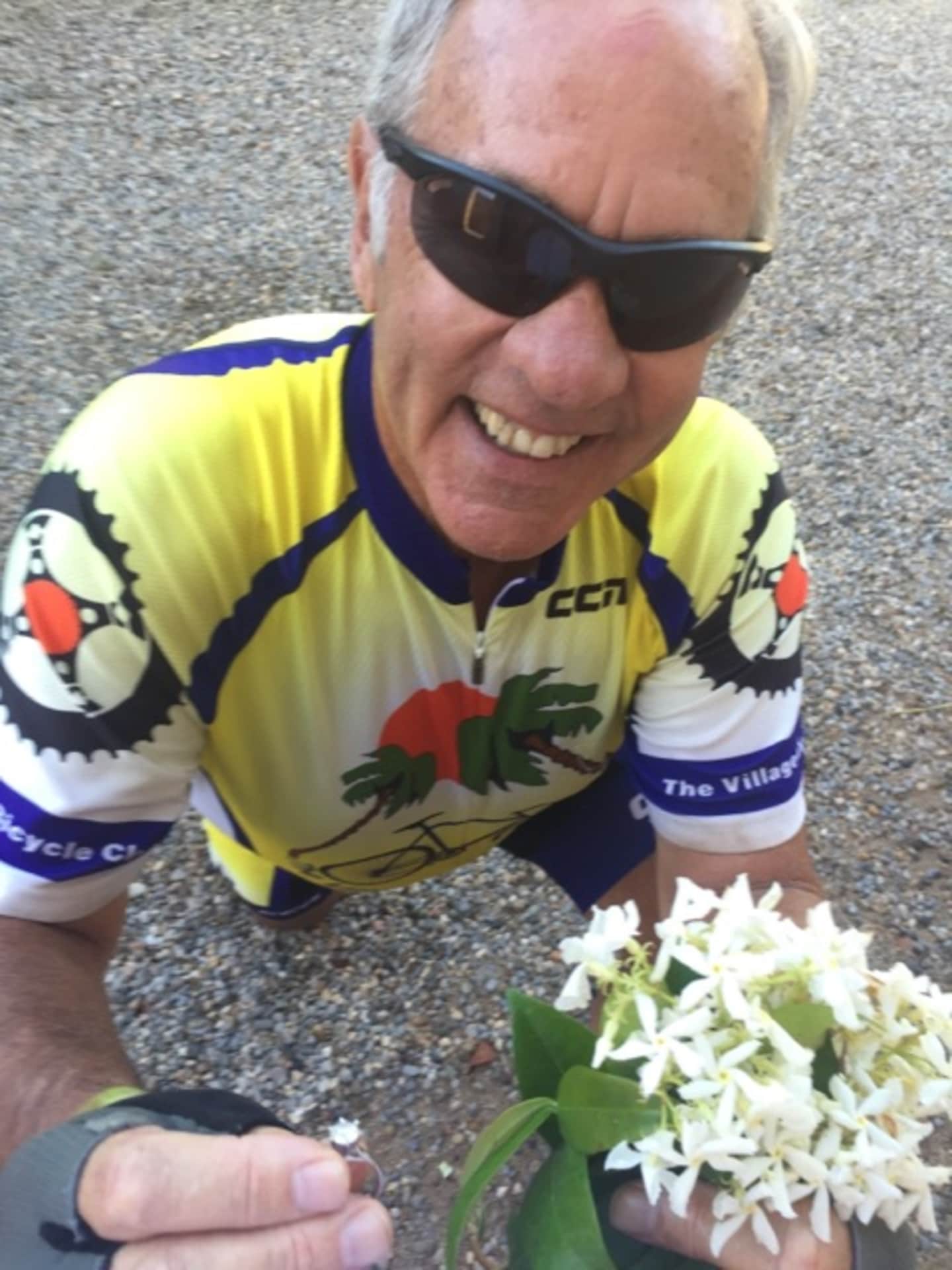 A smiling older man wearing a bright yellow cycling jersey and sunglasses is examining a bouquet of white flowers in the foreground, with a gravel road visible in the background.