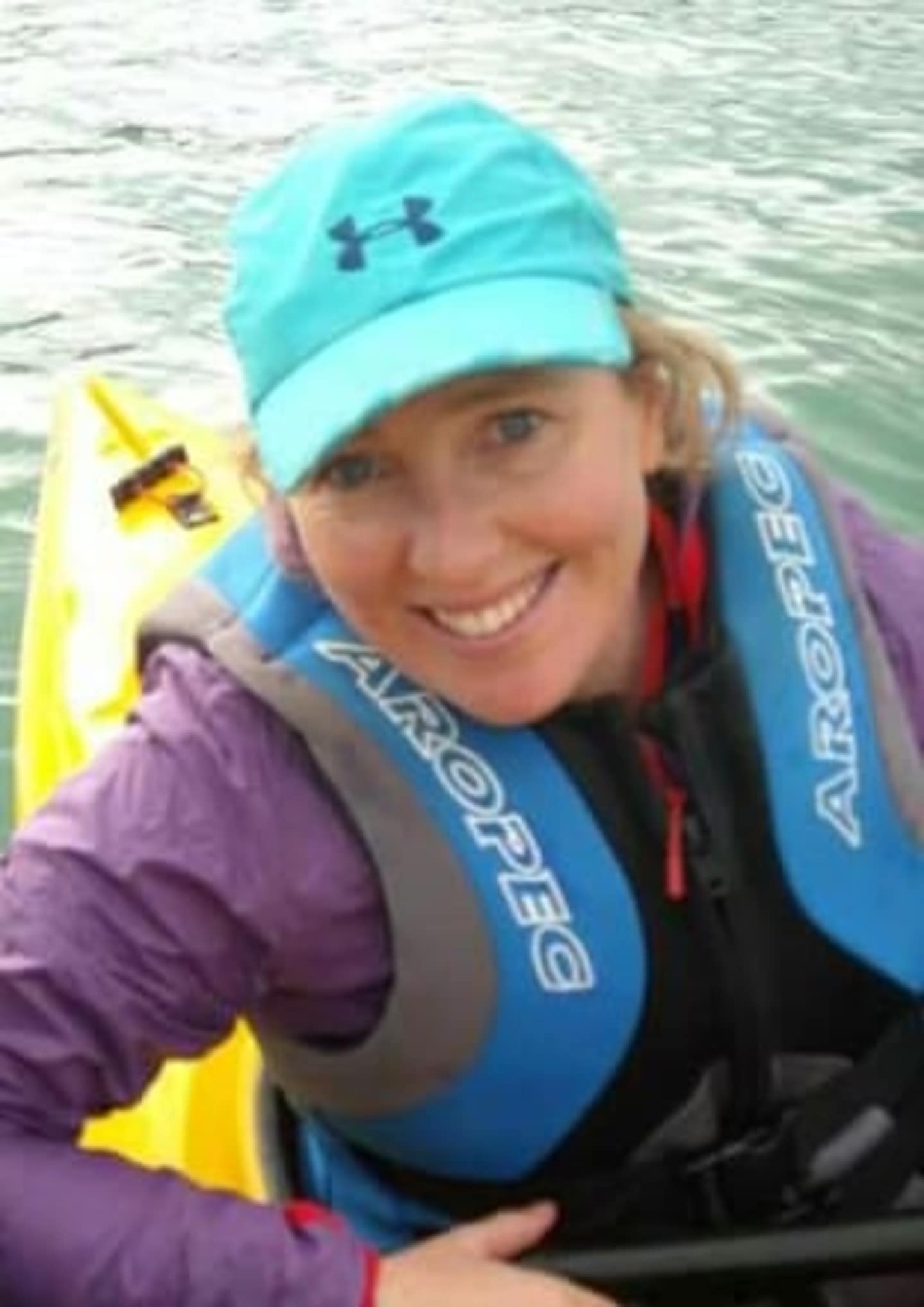 A smiling woman wearing a blue hat and jacket is holding a yellow kayak paddle in the foreground, with a body of water visible in the background.