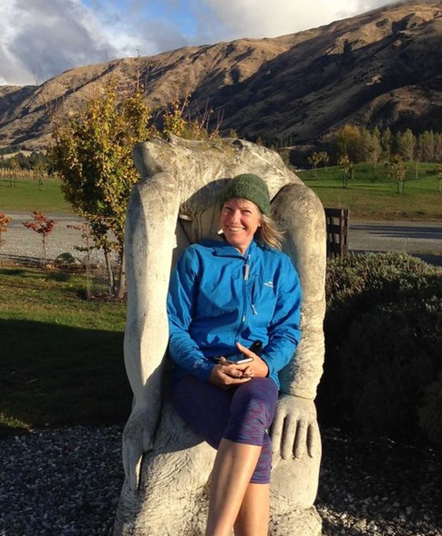 A woman in a blue jacket sits comfortably in a large stone sculpture, surrounded by a scenic landscape of mountains, trees, and a grassy field.
