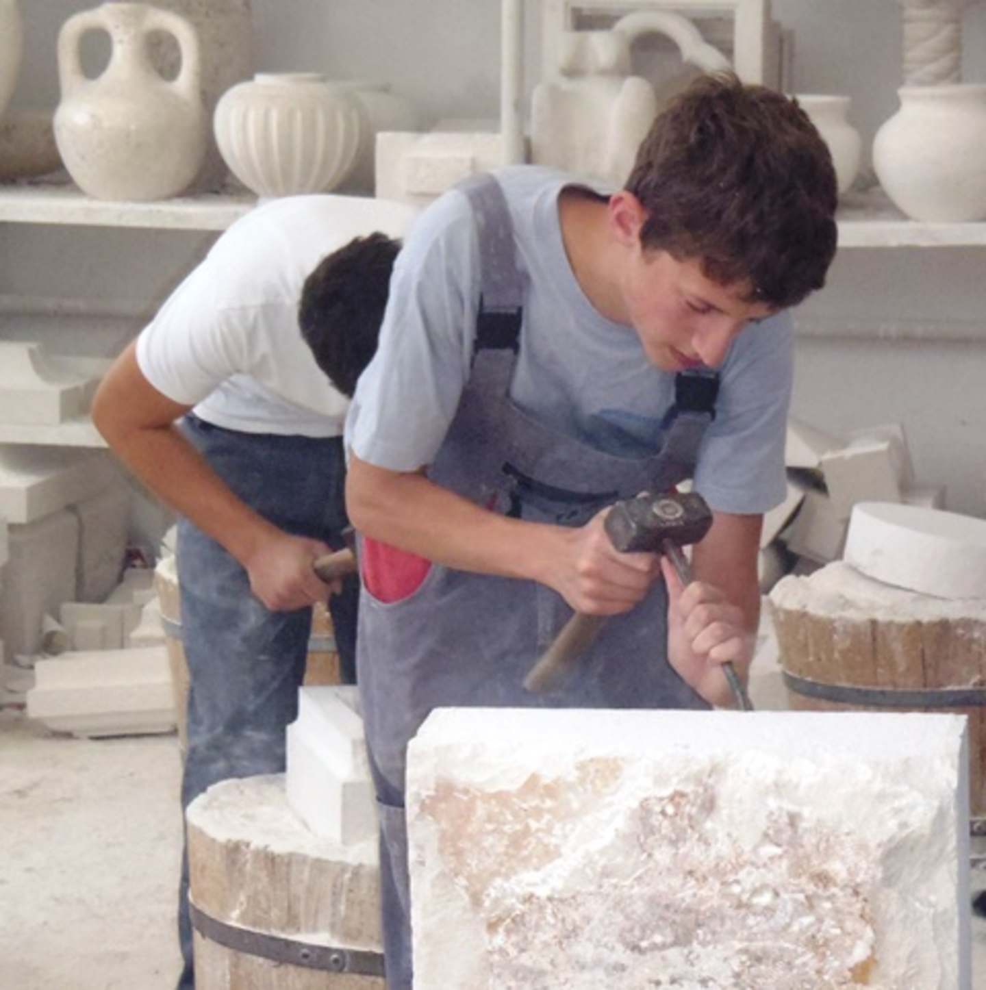 A young man in casual clothing is intently working on carving or sculpting an object on a workbench, surrounded by various ceramic and pottery pieces in the background.