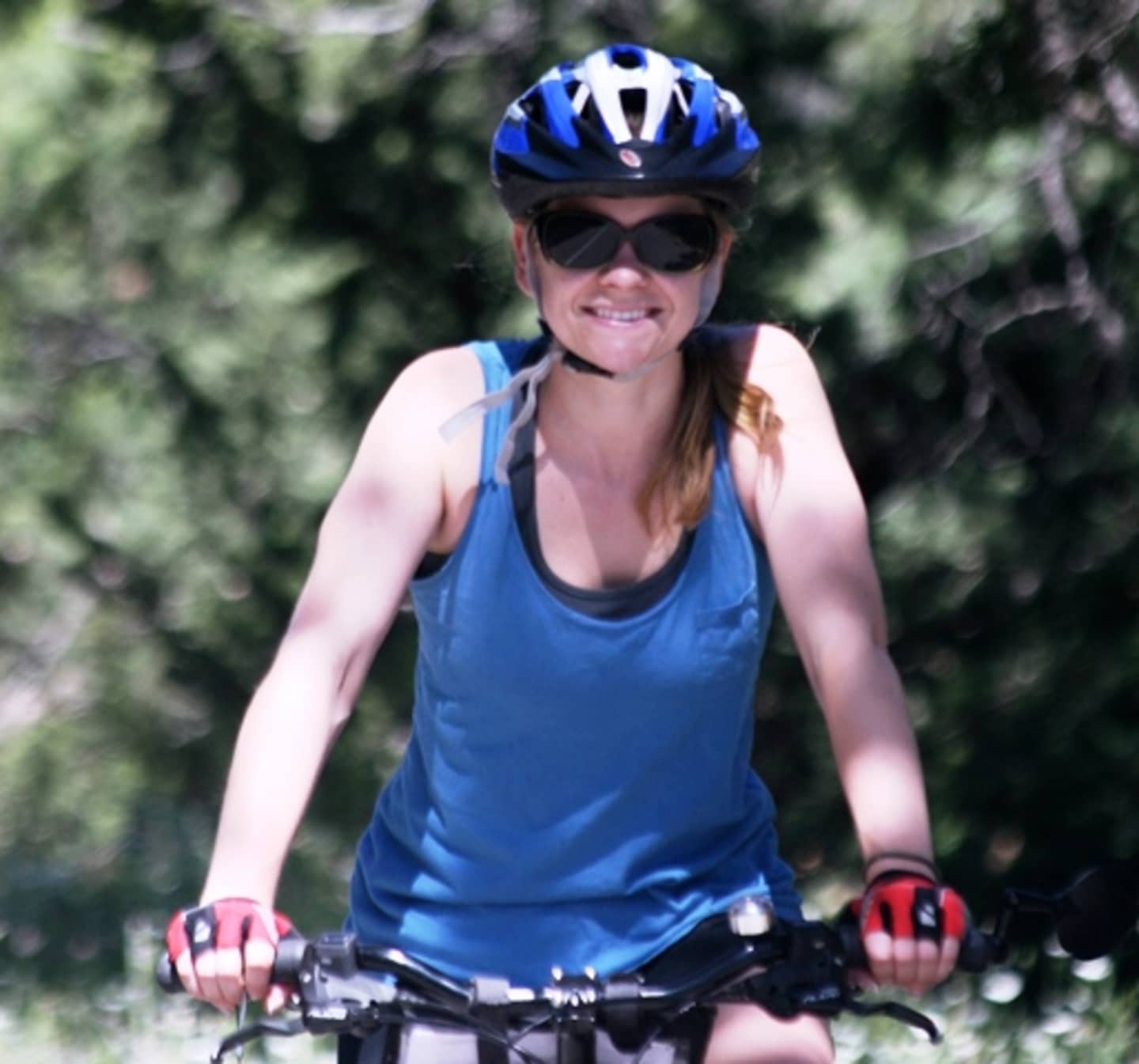A woman wearing a blue tank top, bike helmet, and gloves is riding a bicycle on a wooded trail with lush greenery in the background.