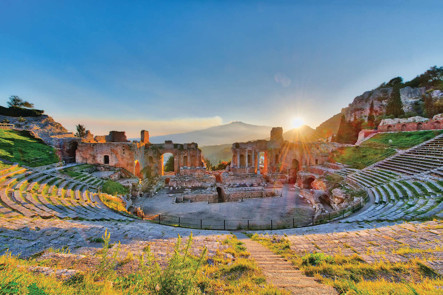 A picturesque ancient Greek amphitheater nestled among rugged mountains, with the sun setting in the distance, casting a warm glow over the historic ruins.