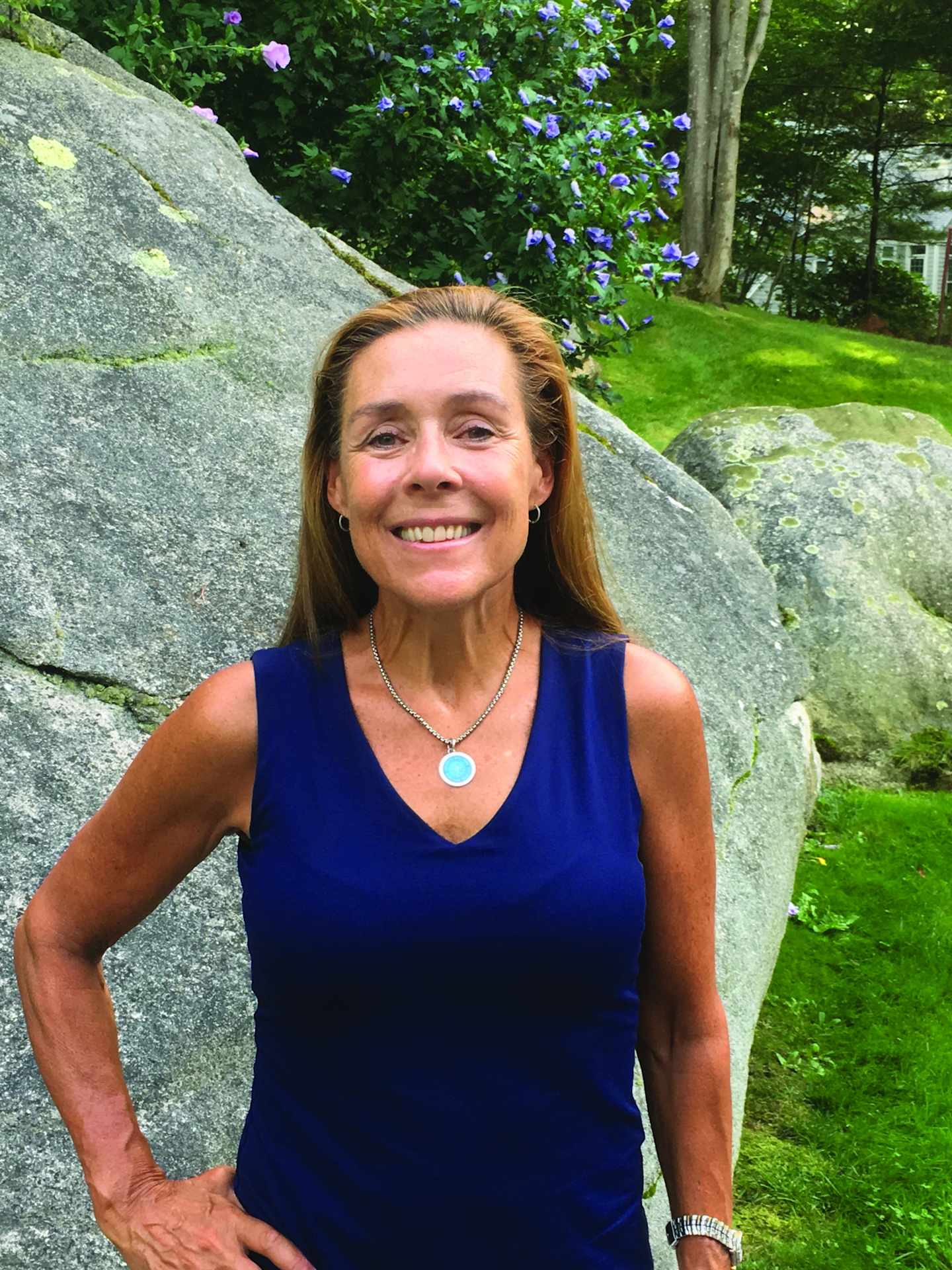 A smiling woman with long brown hair stands in front of a large rock, surrounded by lush green foliage and a grassy lawn in the background.