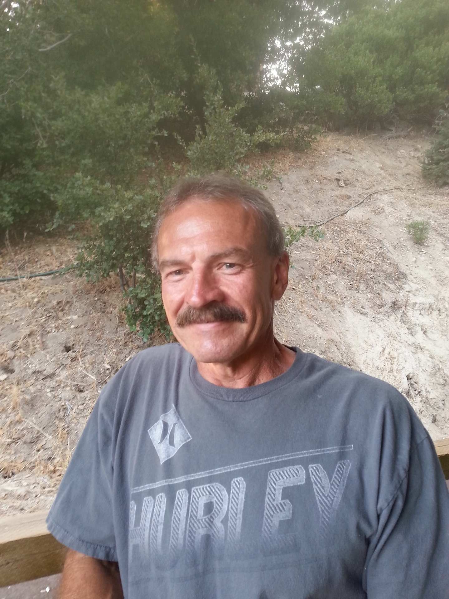 A middle-aged man with a mustache and gray hair is standing in a wooded area, wearing a gray t-shirt with the word "Hurley" printed on it.