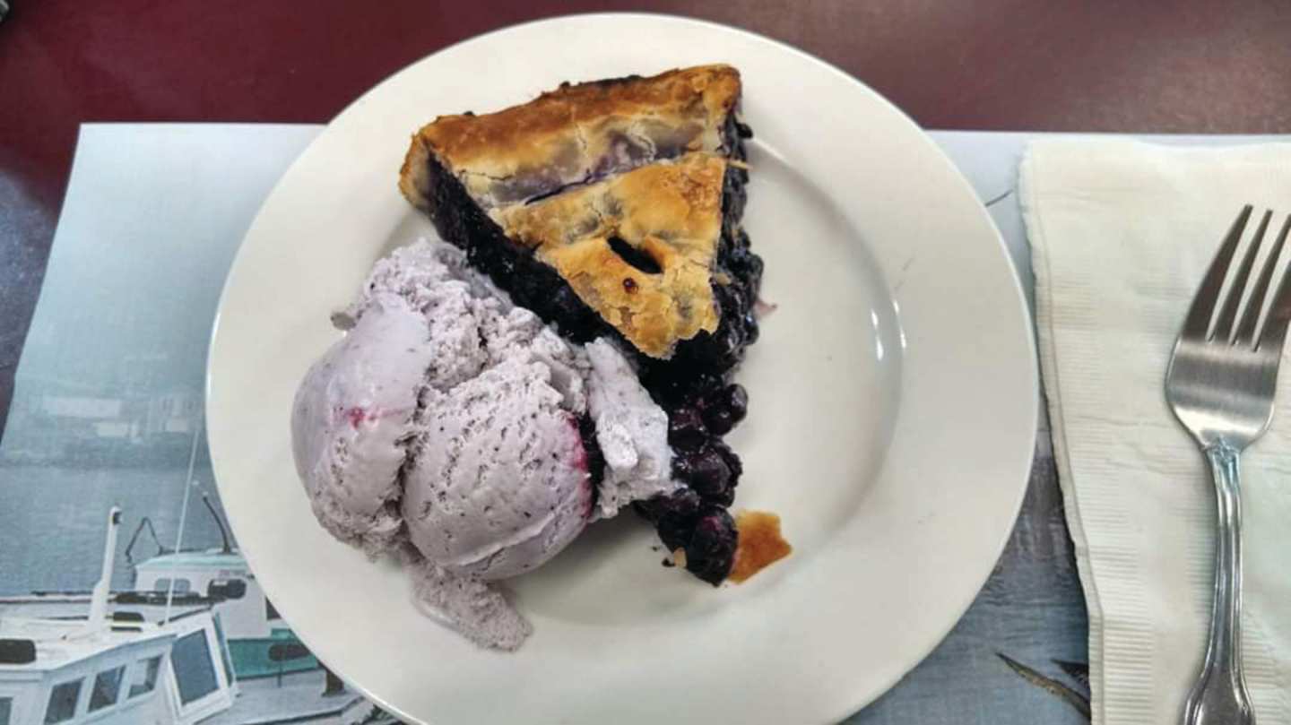 A white plate holds a slice of pie with a scoop of purple-colored ice cream, set against a blurred background that appears to be a window or glass panel.