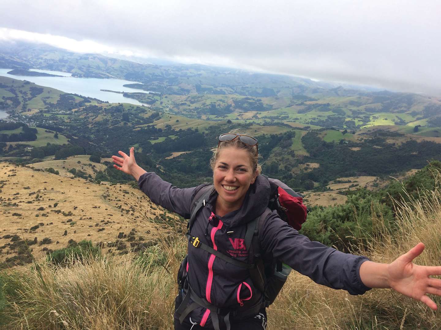 A smiling woman in a hiking jacket stands on a grassy hill, with a scenic landscape of mountains and a lake visible in the background.