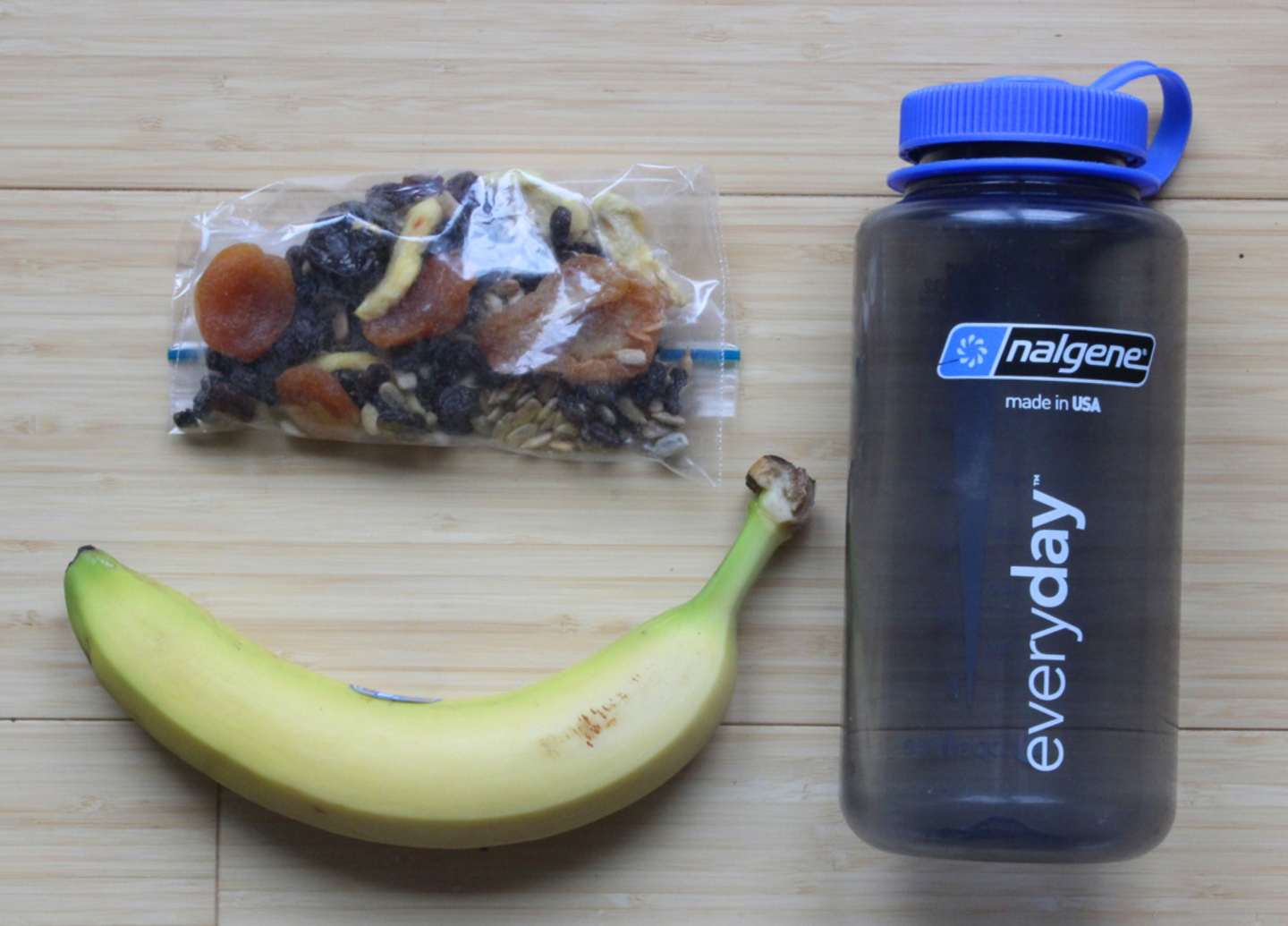 A blue Nalgene water bottle, a plastic bag containing various dried fruits and nuts, and a ripe banana are arranged on a wooden surface.