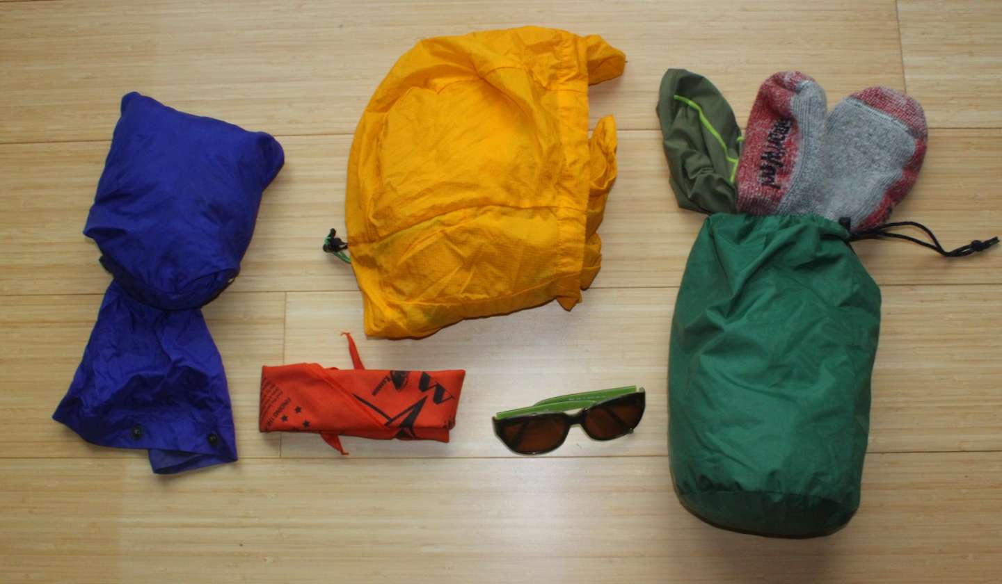 The image shows various outdoor gear items, including a yellow jacket, a blue jacket, a green jacket, a pair of red gloves, and a pair of sunglasses, all arranged on a wooden surface.
