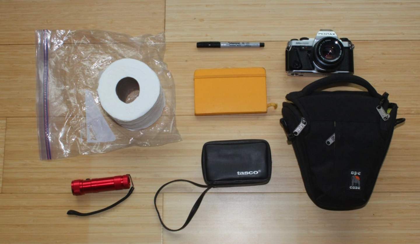 The image shows various office and photography-related items on a wooden surface, including a camera, a power bank, a notebook, and a roll of toilet paper.