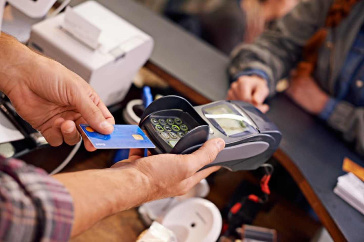 A person is using a credit card to make a payment at a payment terminal, with other payment devices and a cash register visible in the background.