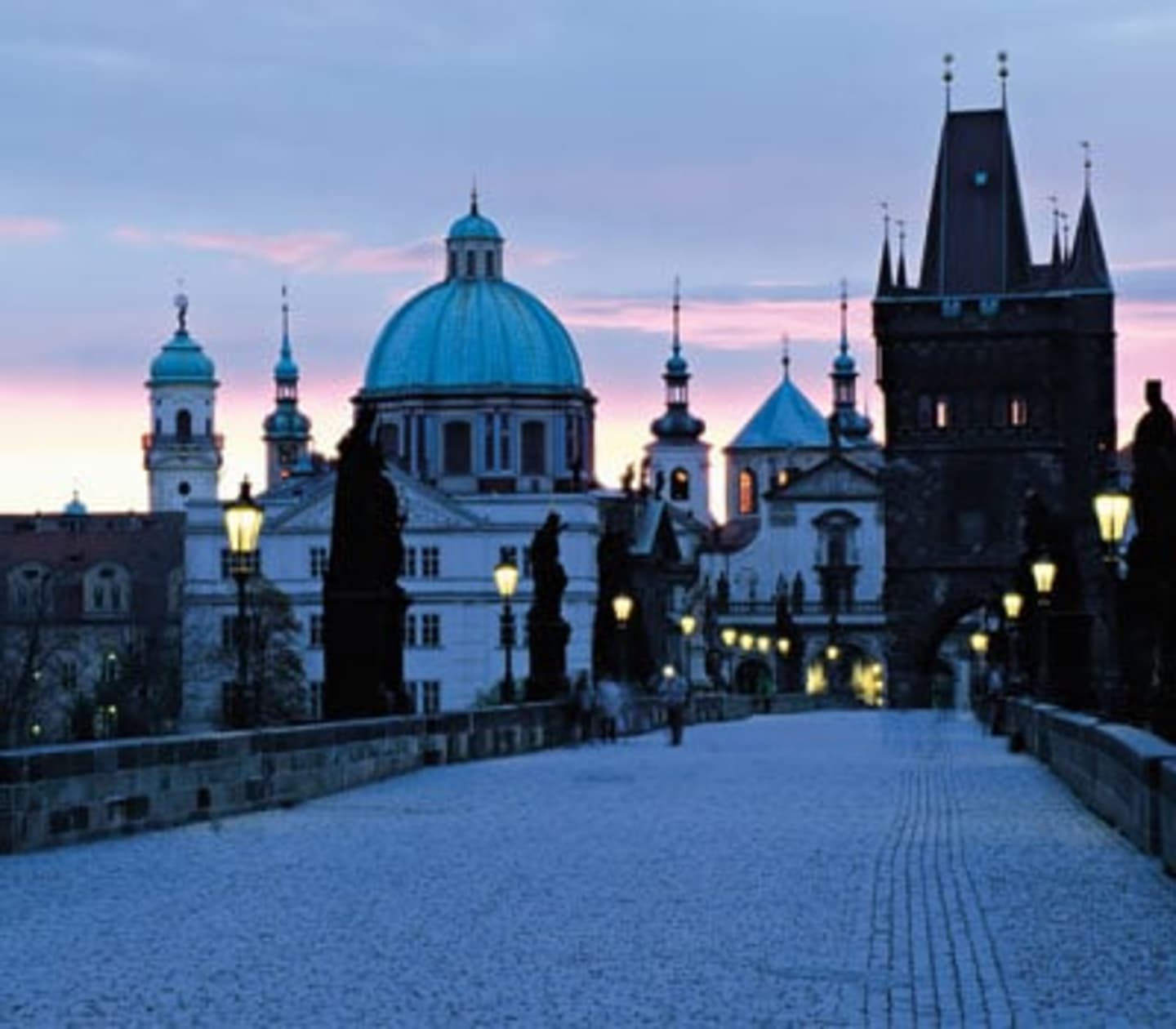 A snow-covered bridge leads to a picturesque cityscape with ornate domes, spires, and towers against a colorful evening sky.