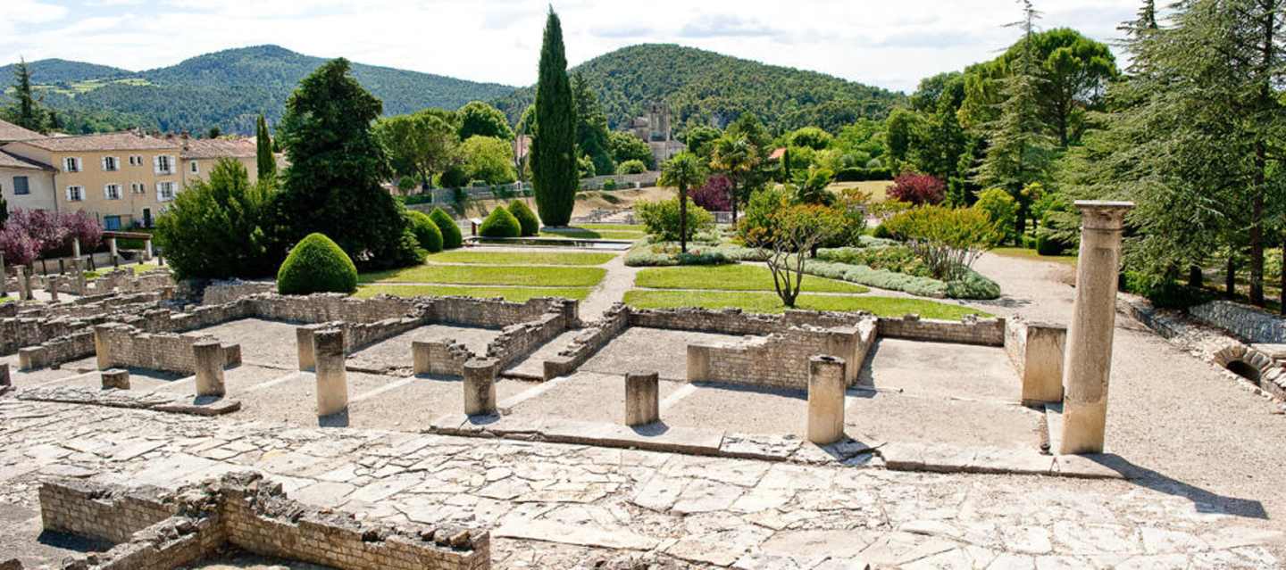 A well-manicured garden with neatly trimmed hedges and trees, surrounded by ancient stone ruins and lush green mountains in the background.
