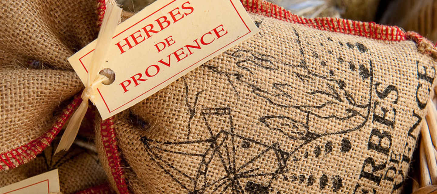 The image shows a burlap sack or bag with a label that reads "HERBES DE PROVENCE" in red text, and the background appears to be a woven or textured surface.