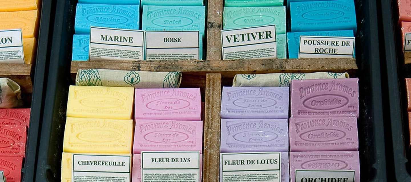 The image shows various boxes of different scented soaps, including marine, boise, vetiver, poussiere de roche, chevre feuille, fleur de lis, fleur de lotus, and orchidee, arranged on wooden shelves.