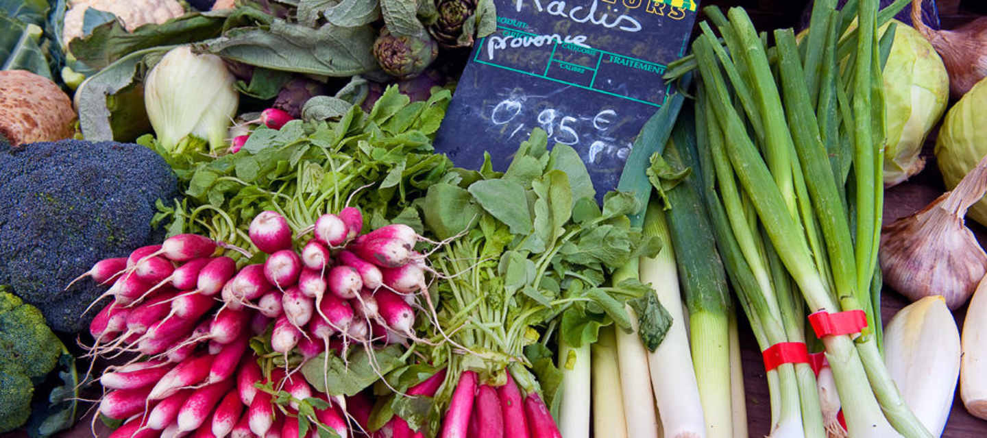 A vibrant display of fresh produce, including bunches of radishes, leafy greens, and green onions, arranged on a market stall with a chalkboard sign displaying prices.