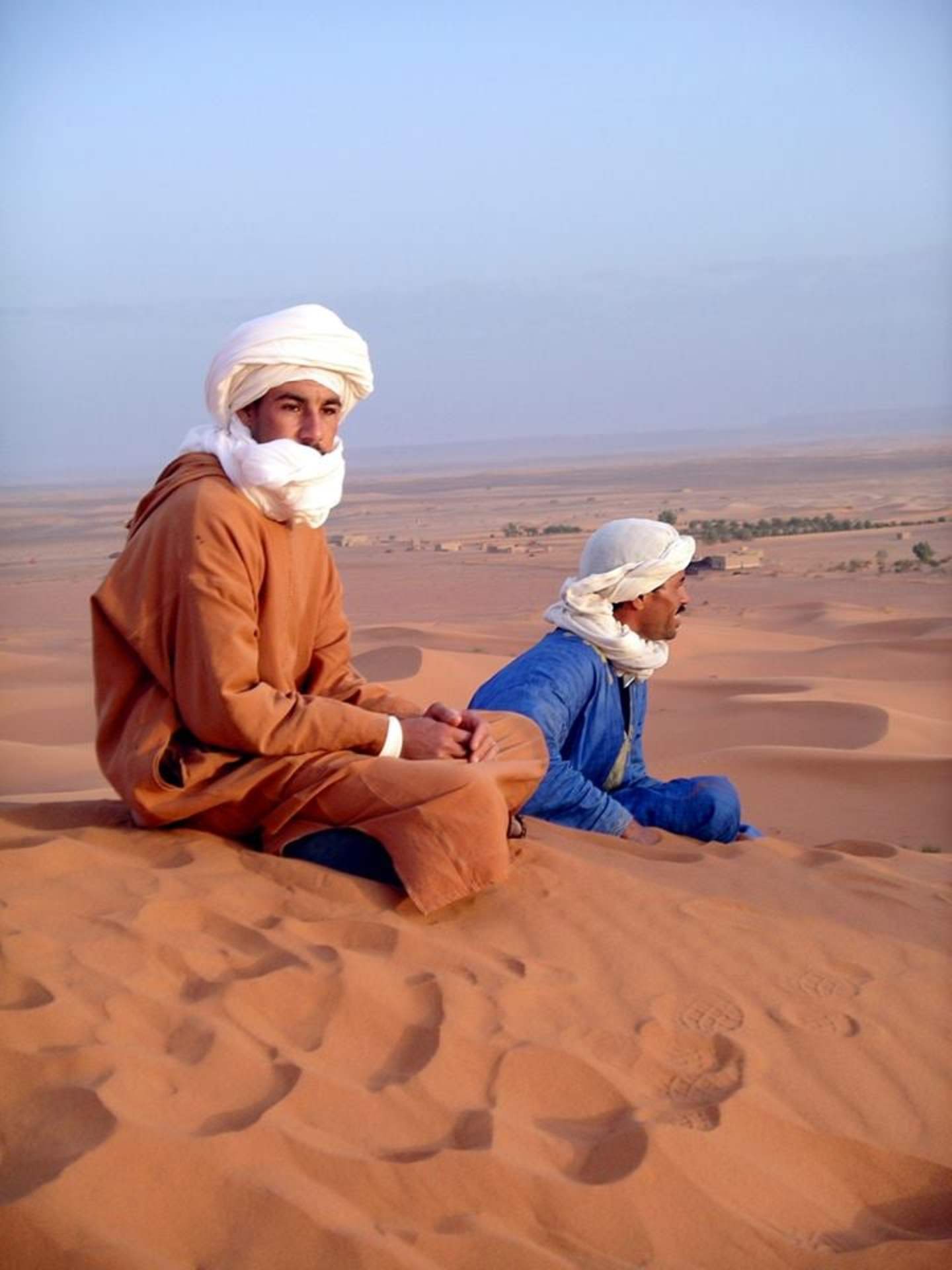 Two individuals, dressed in traditional desert attire, sit on the sand in a vast, arid landscape with a clear blue sky in the background.