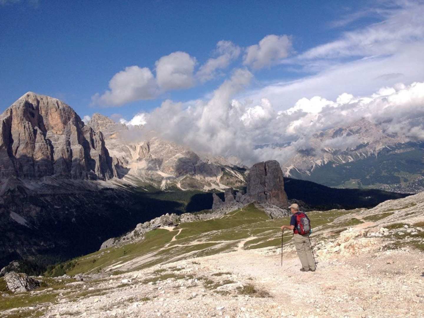 A hiker stands on a rocky trail in the foreground, surrounded by towering, rugged mountains and dramatic clouds in the background.
