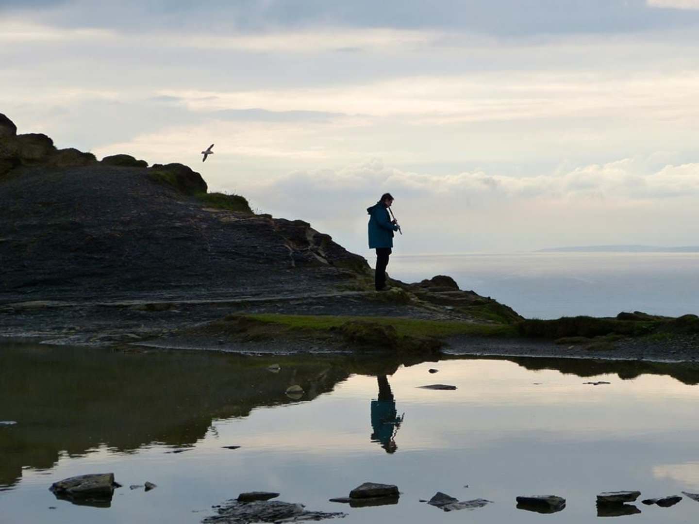 A lone figure stands on a rocky outcrop, reflected in the still waters below, with a bird soaring overhead against a cloudy sky.