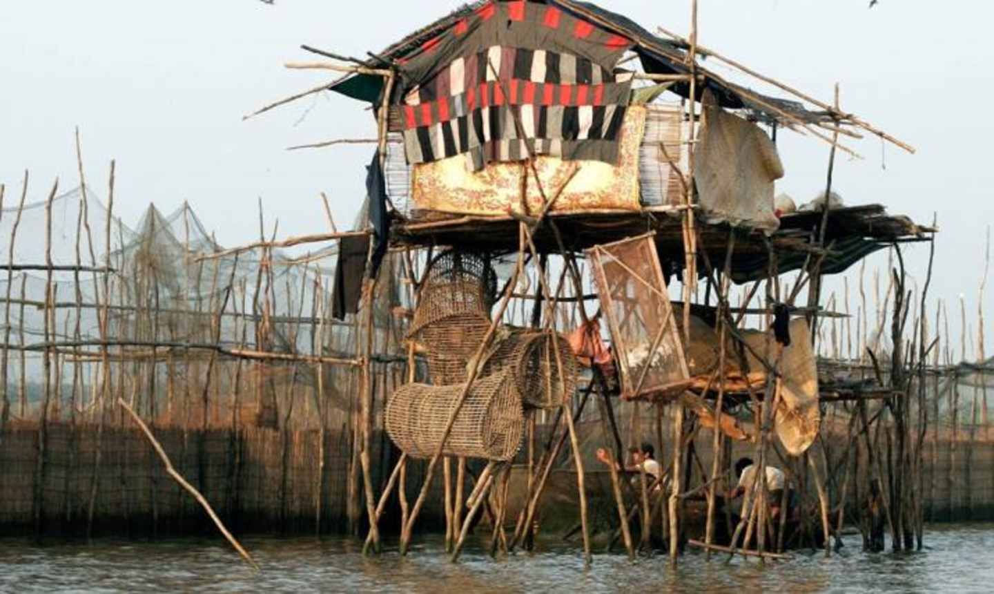 A colorful and intricate structure made of bamboo and other natural materials stands in a body of water, surrounded by tall reeds and a hazy background.