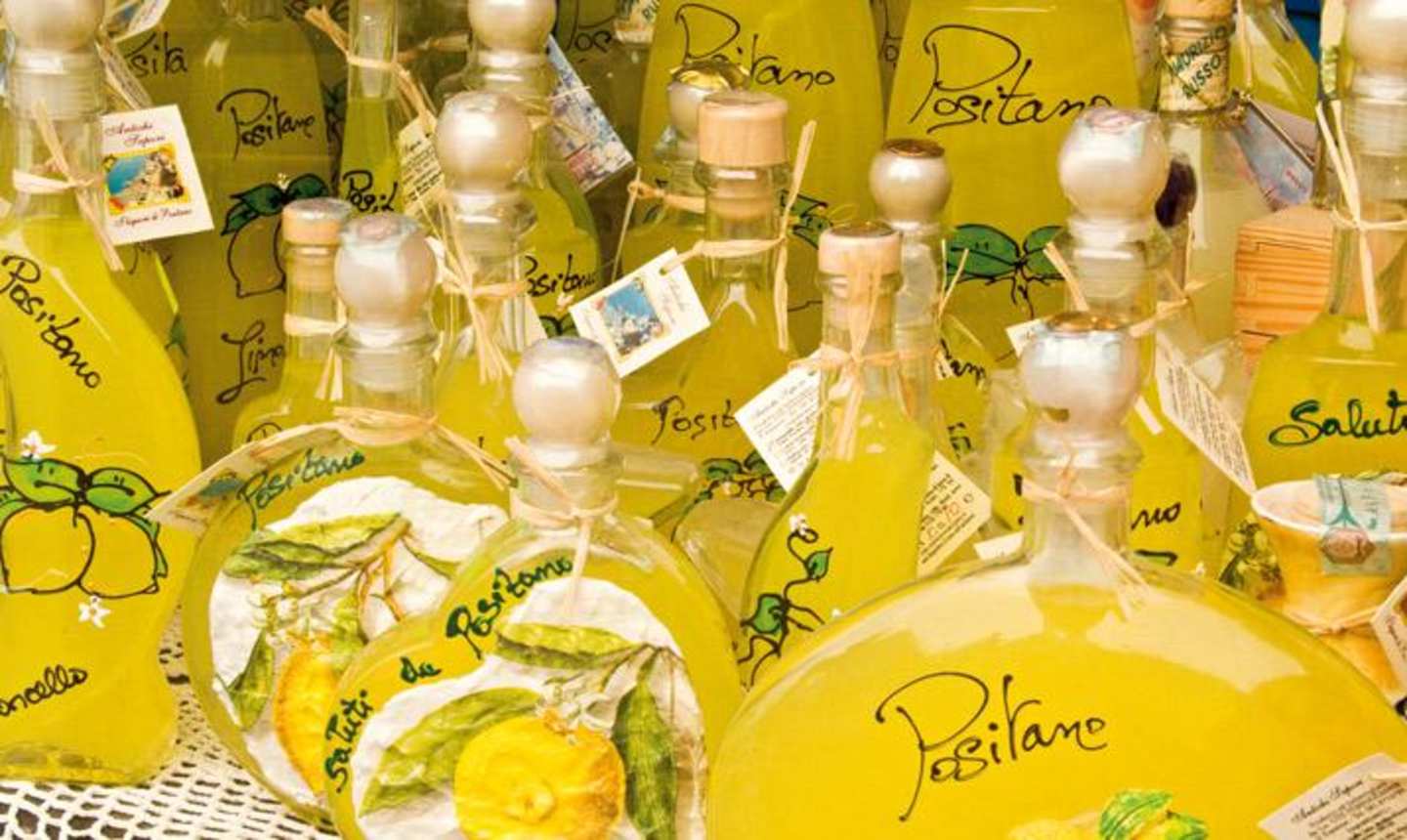 Numerous bottles filled with a yellow liquid, likely olive oil, are displayed with handwritten labels and decorative elements, creating a vibrant and visually appealing arrangement.