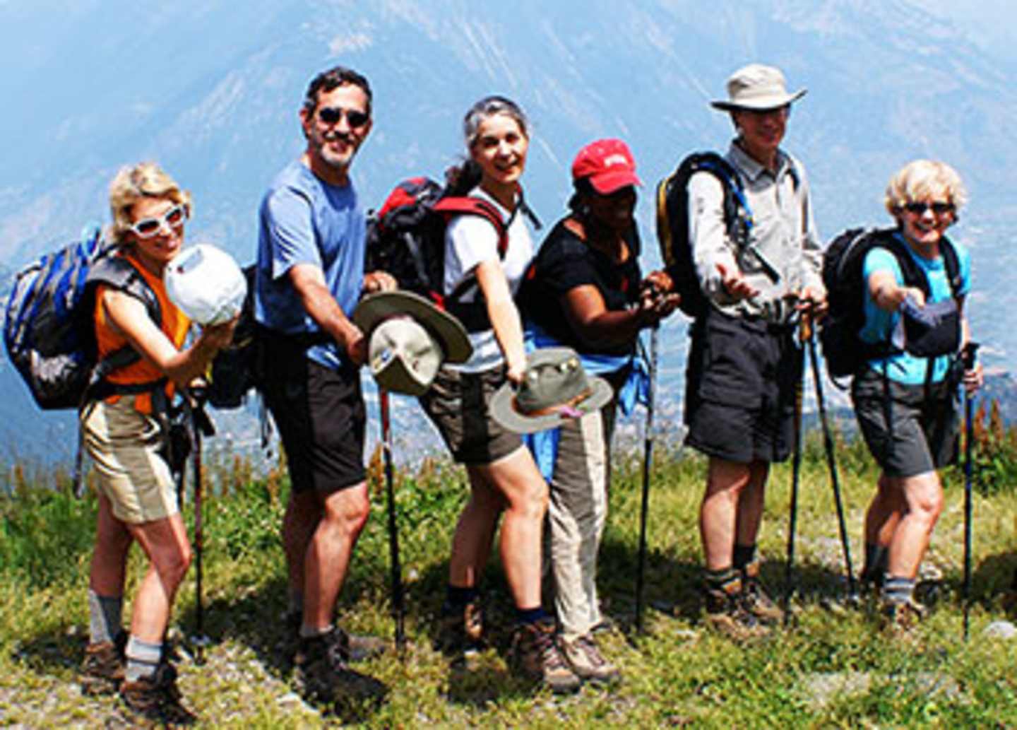 A group of hikers, dressed in outdoor gear, stands together in a grassy field with mountains visible in the background.