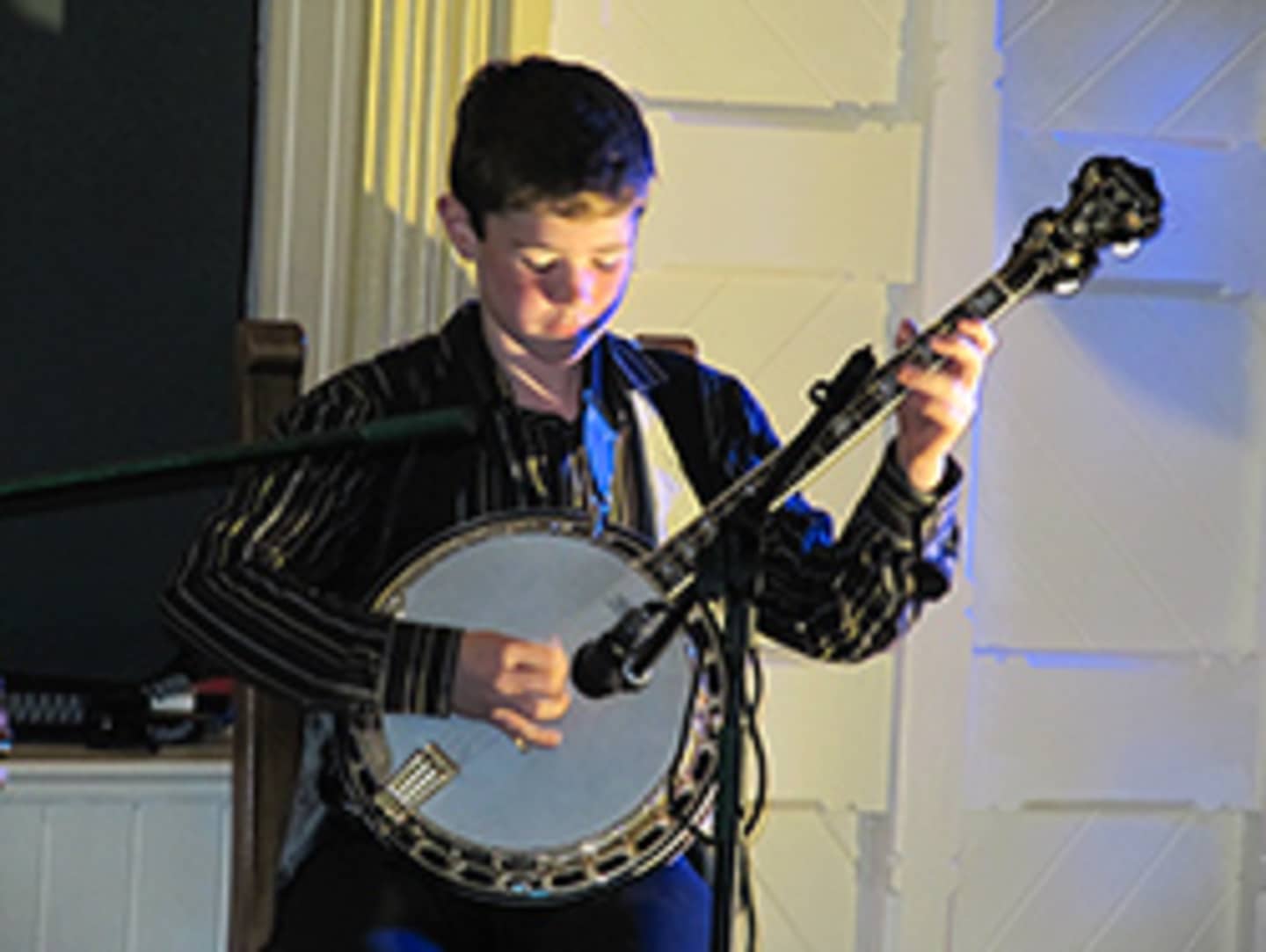 A young person is playing a banjo in a dimly lit room with curtains in the background.