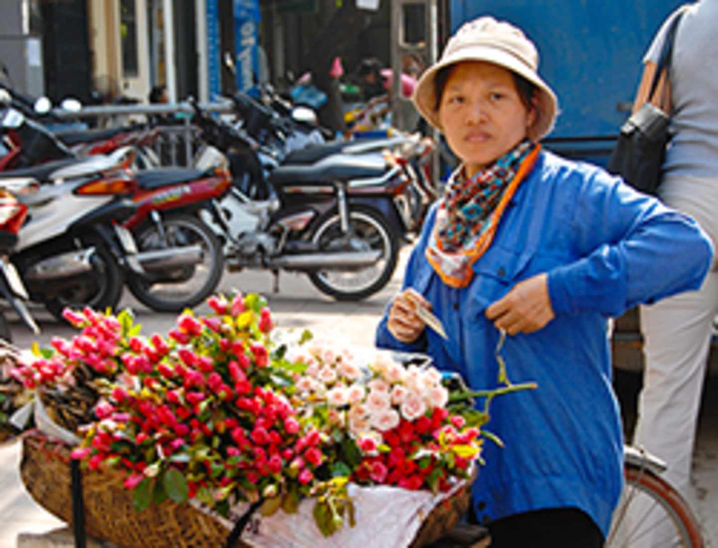 A smiling elderly woman in a blue jacket and hat stands in front of a display of vibrant red and pink flowers, with a backdrop of parked motorcycles.