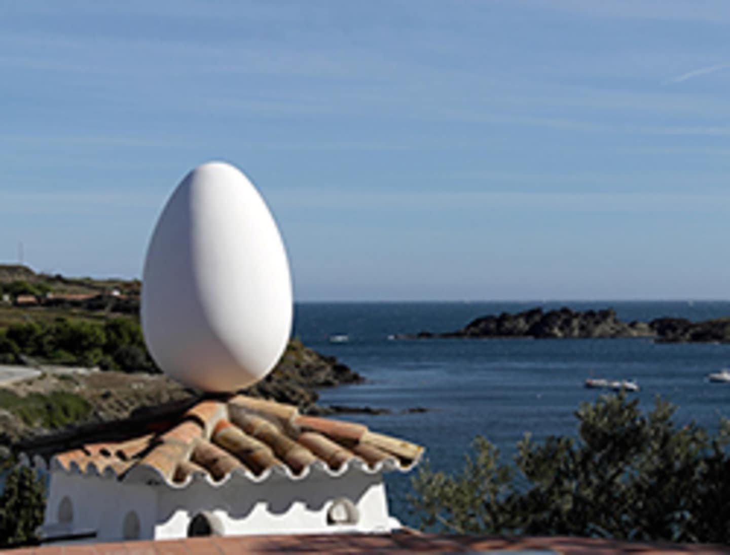 A large, white, egg-shaped sculpture stands on a pedestal against the backdrop of a scenic coastal landscape with rocky cliffs and a calm blue ocean.