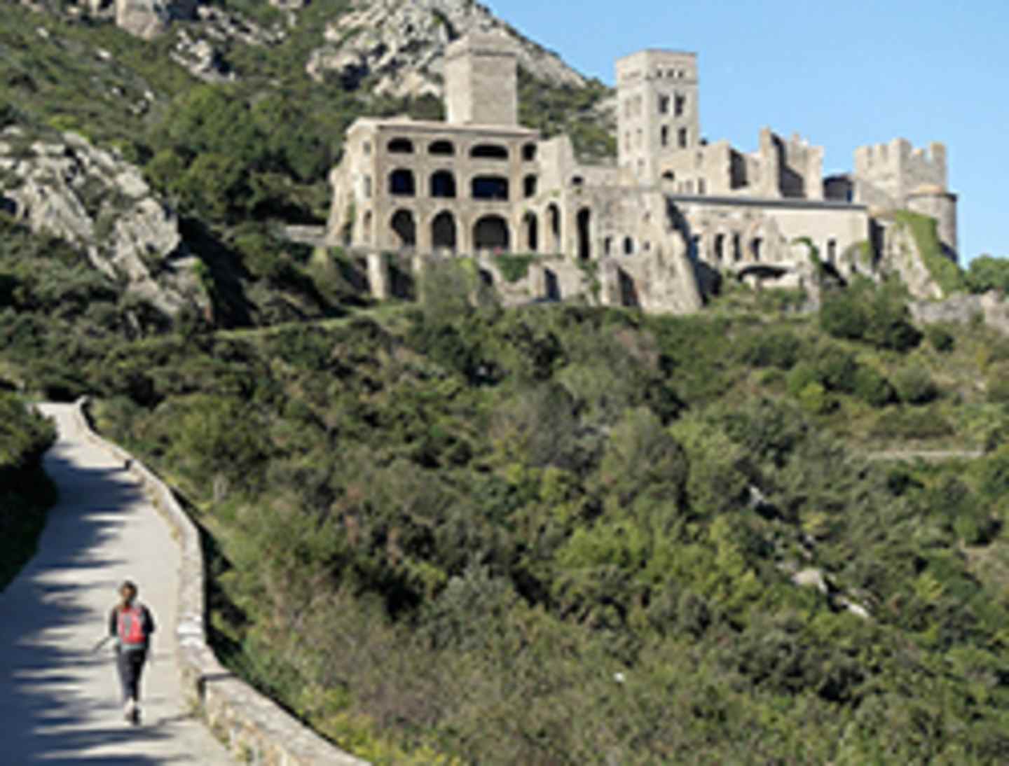 A winding path leads through lush greenery towards an impressive medieval-style castle perched atop a rocky outcrop, its towers and turrets rising majestically against the clear blue sky.