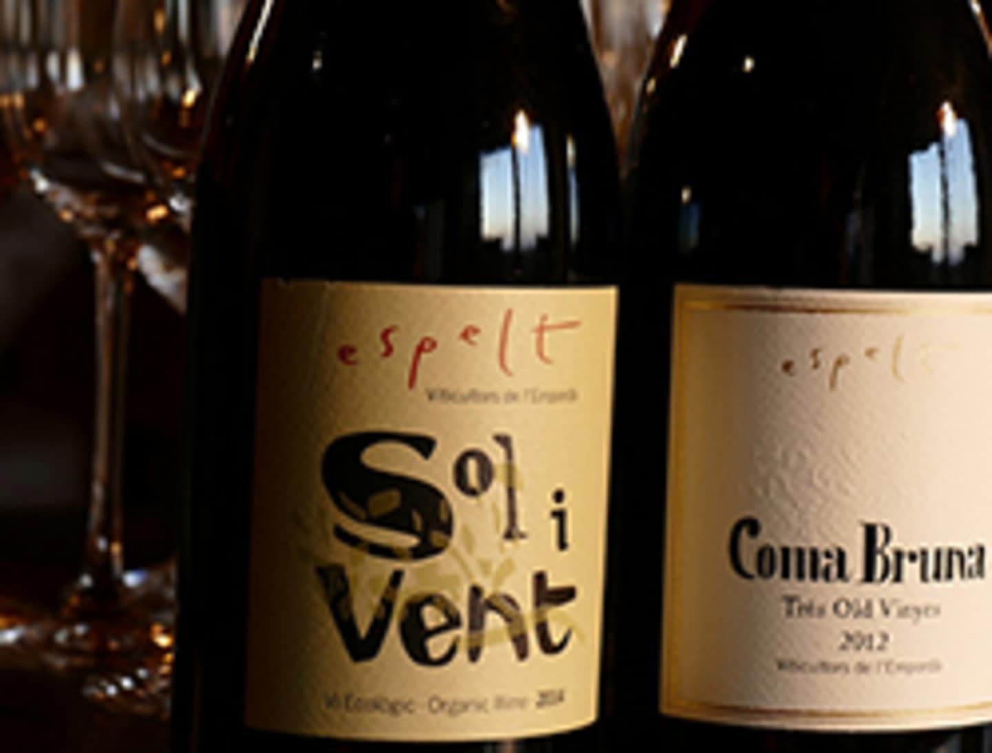 The image shows two bottles of wine, one labeled "Sol i Vent" and the other "Coma Bruna", set against a blurred background of wine glasses.