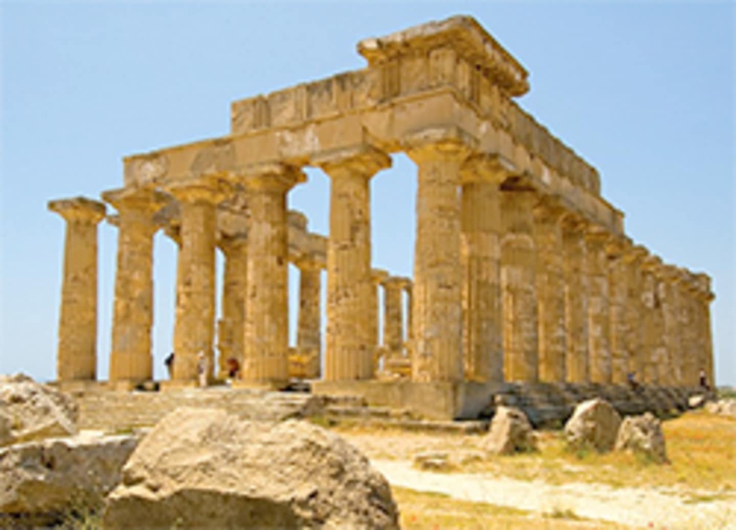 The image depicts the ancient ruins of a Greek temple, with its iconic columns and pediment standing tall against a clear blue sky. In the foreground, there are several large rocks and debris, adding to the sense of the site's historical significance.