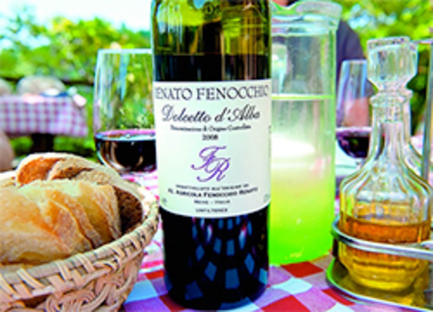 A bottle of Exato Fenocchio Dolcetto d'Alba wine surrounded by a basket of bread, glasses, and a colorful tablecloth in an outdoor setting with greenery in the background.