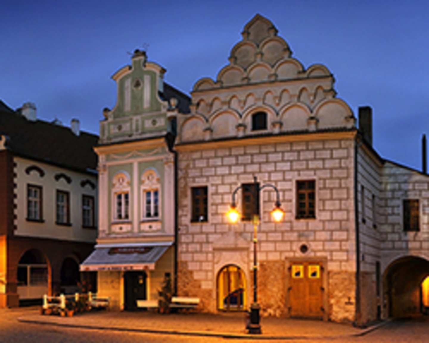 A historic, ornately decorated building with arched windows and a distinctive roofline stands in the foreground, surrounded by other buildings in a quaint, dimly lit town setting.