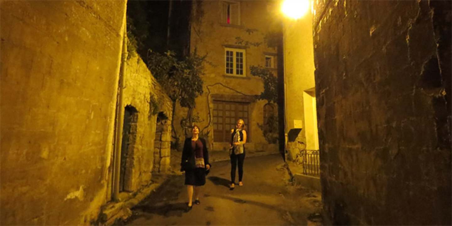 A dimly lit alleyway with two people walking through it, surrounded by old stone buildings and a warm, golden glow from the lighting.