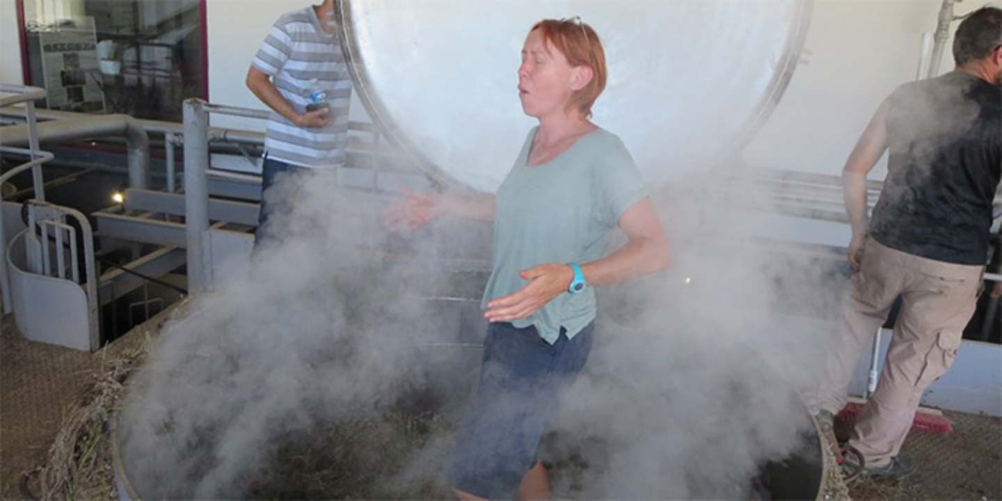 The image shows a person standing in a cloud of dust or smoke, surrounded by other individuals in a workshop or industrial setting.
