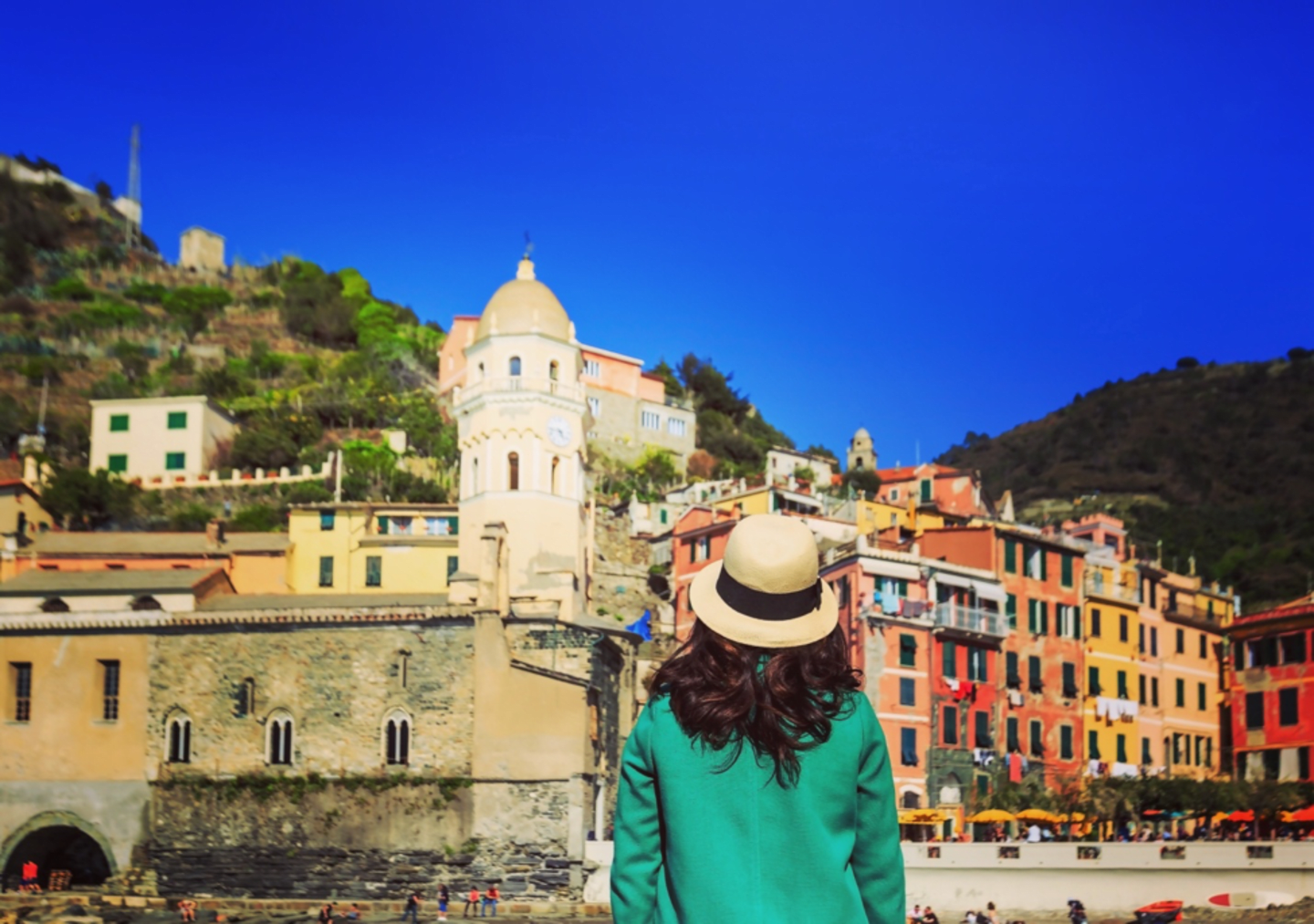 A woman wearing a green sweater and a hat stands in the foreground, facing away from the camera, with a picturesque Italian town nestled in the hills behind her.