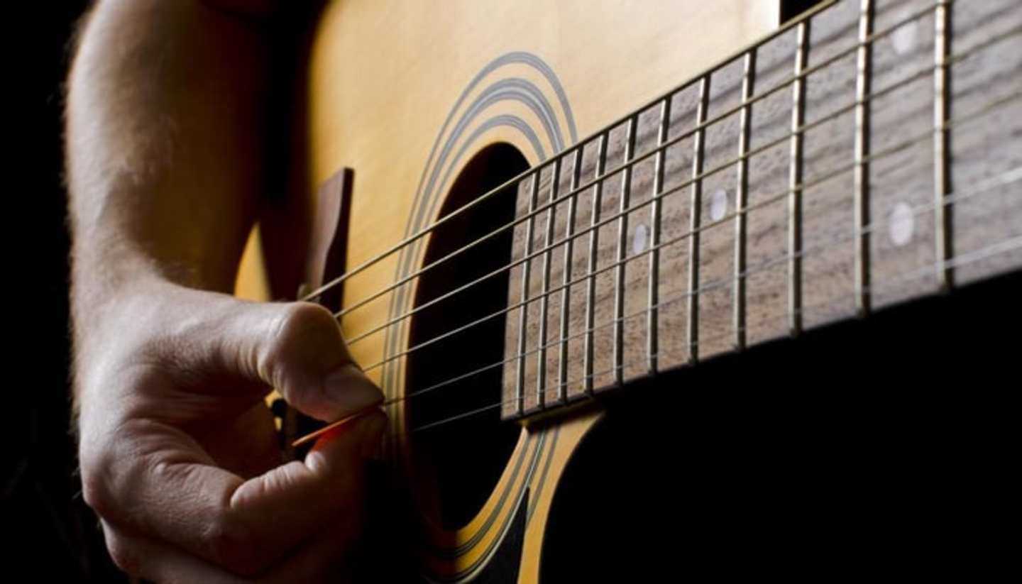 A close-up view of a person's hand playing an acoustic guitar, with the guitar's fretboard and strings prominently featured in the foreground against a dark background.