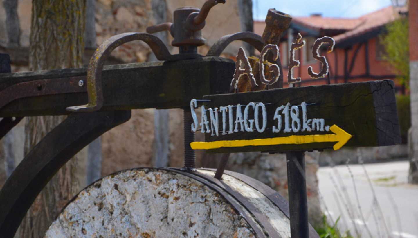 A rustic signpost with the text "Santiago 516km" points the way, set against a backdrop of weathered wooden structures and a stone wall.