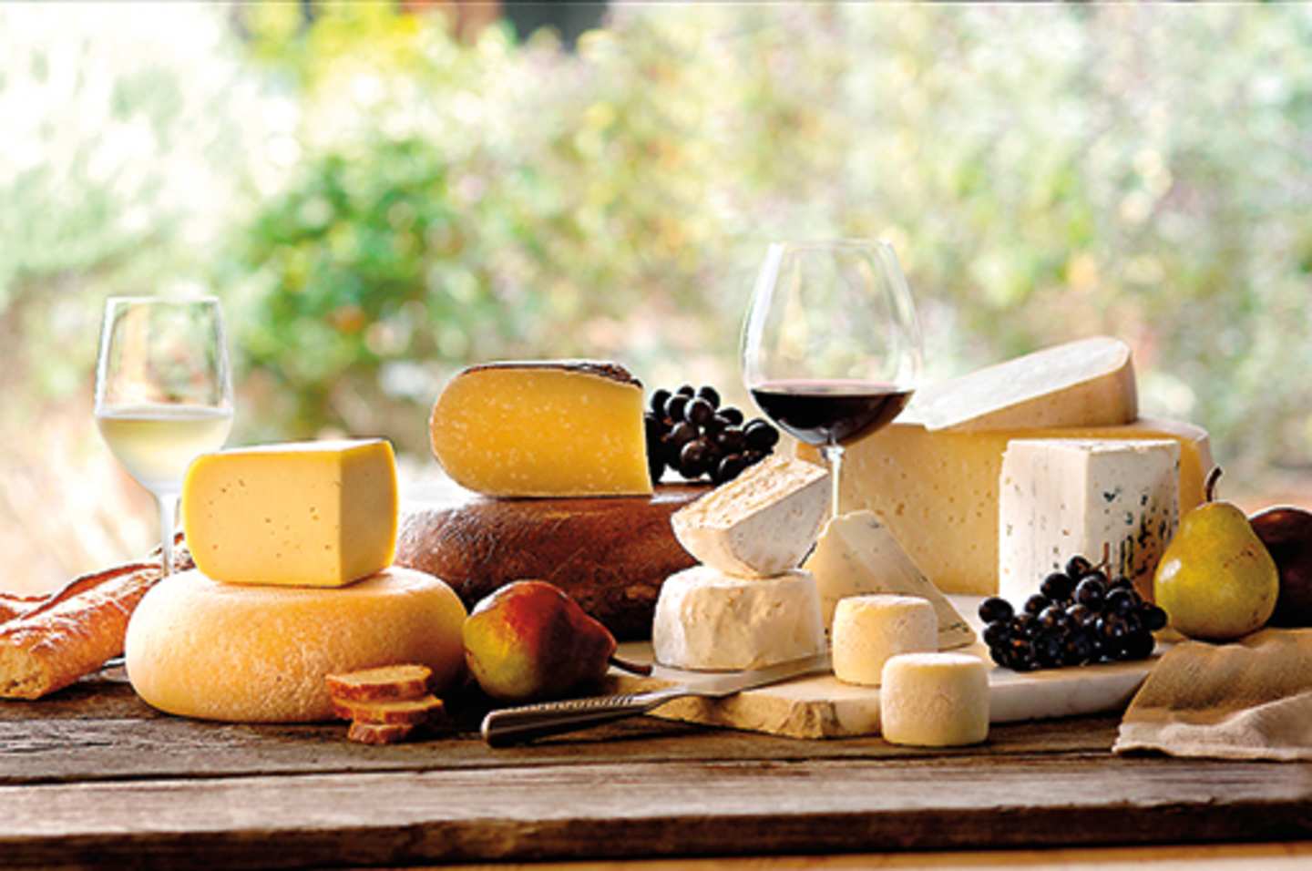 A rustic wooden table displays an assortment of artisanal cheeses, fresh fruits, and a glass of red wine, set against a blurred natural backdrop.