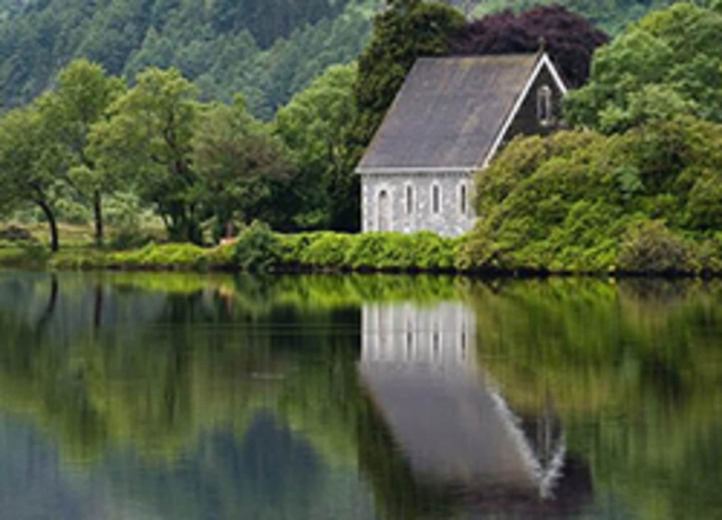 A small wooden church building is reflected in the calm waters of a lake, surrounded by lush green trees and a mountainous landscape in the background.