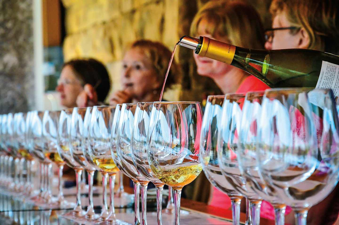 A row of wine glasses filled with amber liquid, with people in the background pouring from a bottle into the glasses.