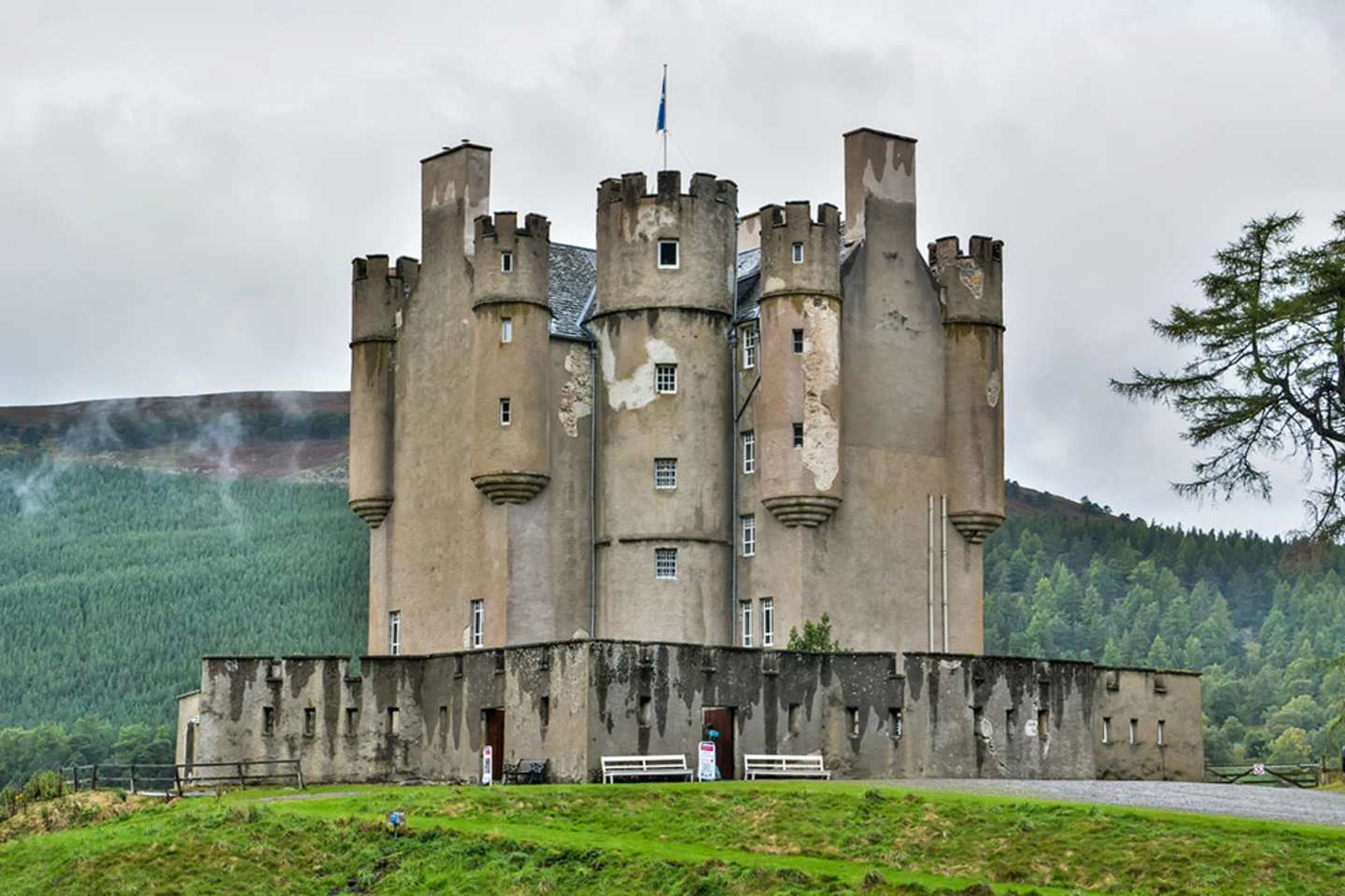 A towering medieval-style castle stands on a grassy hill, surrounded by lush forests and a cloudy sky in the background.