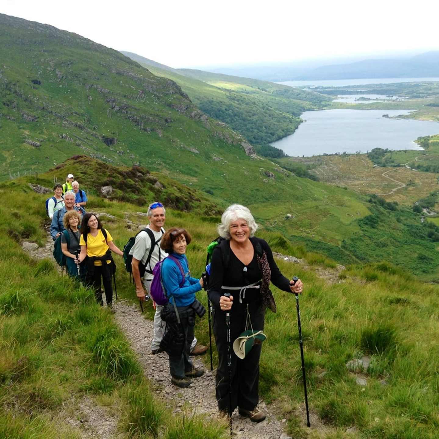 A group of hikers, including an older woman with white hair, are walking on a trail through a lush, green landscape with a lake visible in the background.