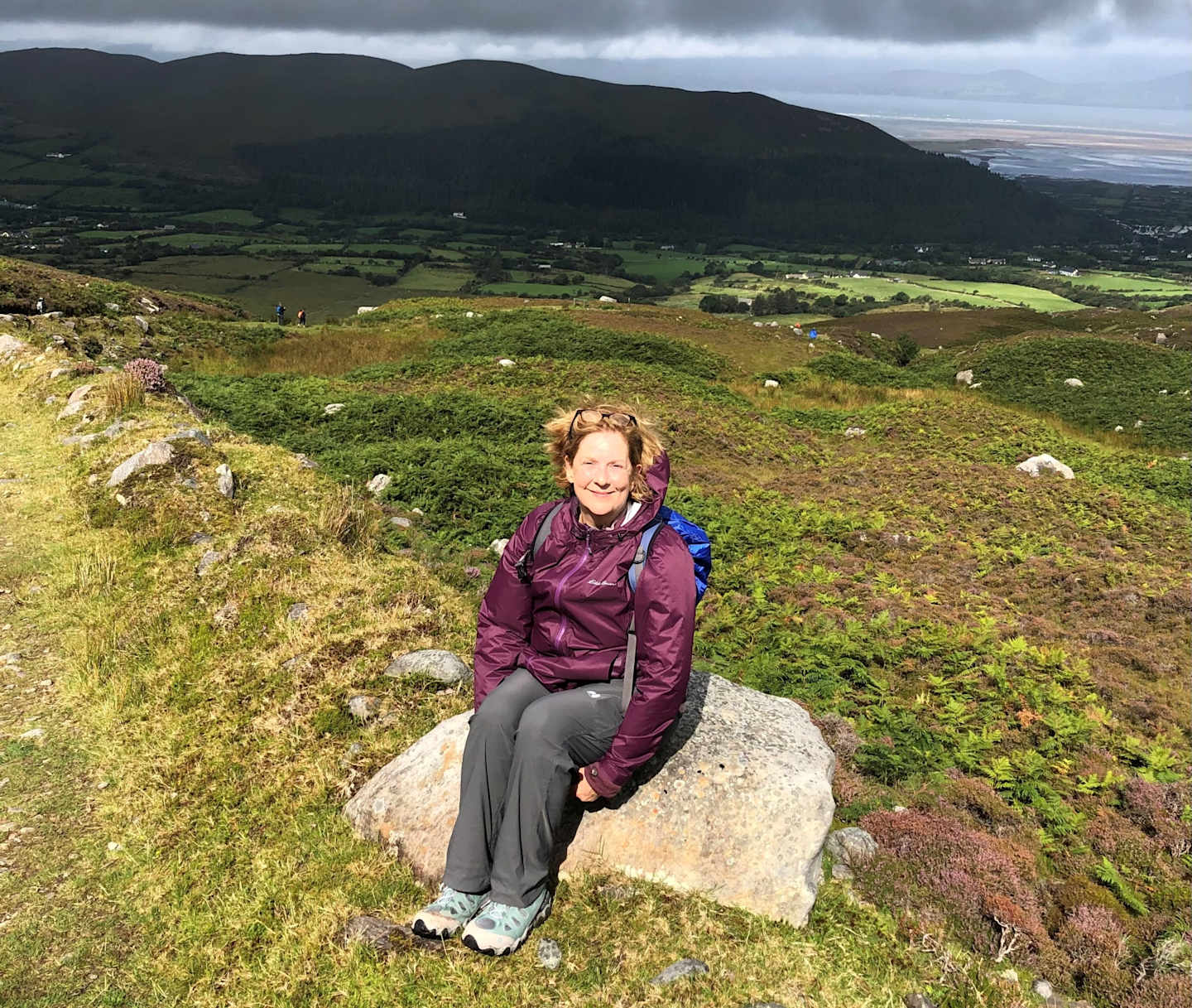 A person sits on a large rock in a grassy field, with mountains and a cloudy sky visible in the background.