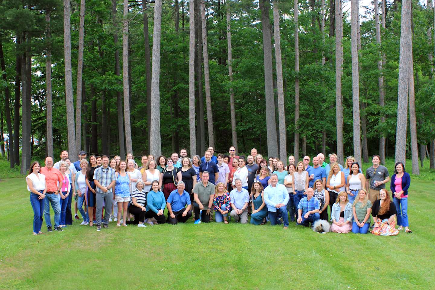 A large group of people, mostly women, gathered together in a grassy area surrounded by a dense forest of tall trees.