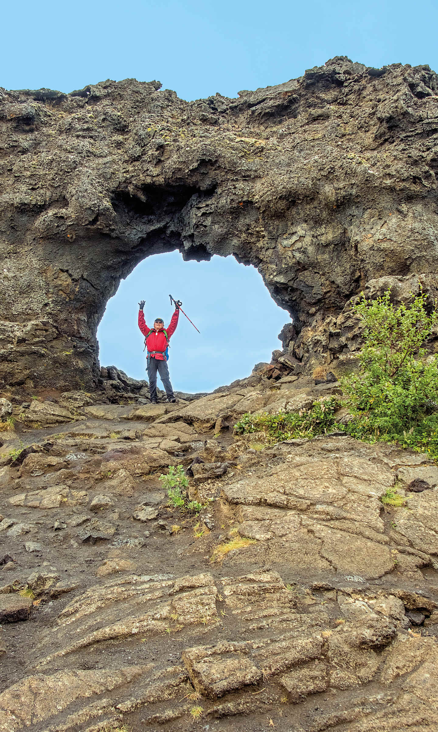 A person in a red jacket stands triumphantly within a natural rock arch, with a rugged, mountainous landscape visible in the background.