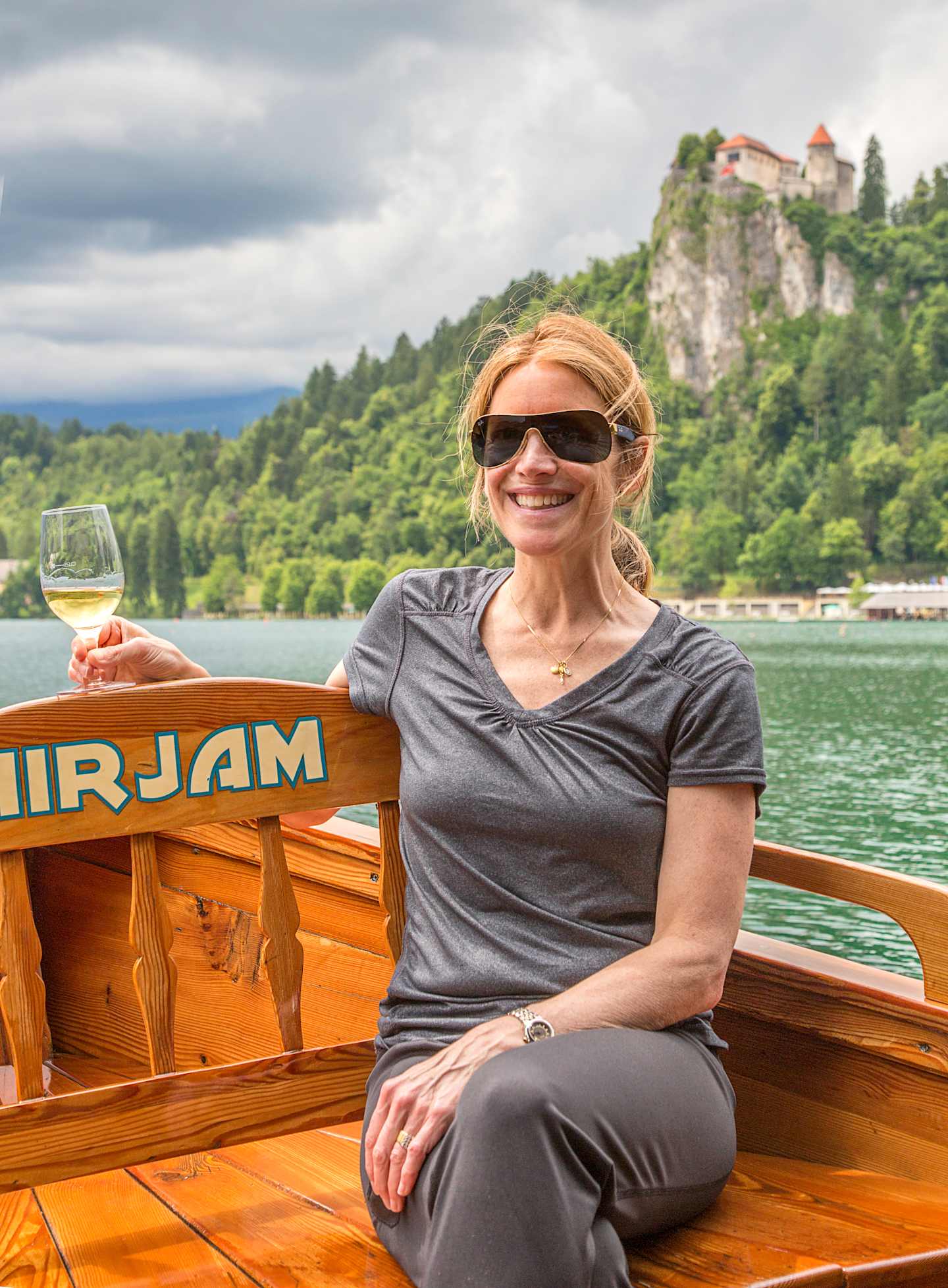 A smiling woman in sunglasses sits in a wooden boat on a lake, with a castle-like structure visible in the background against a cloudy sky.