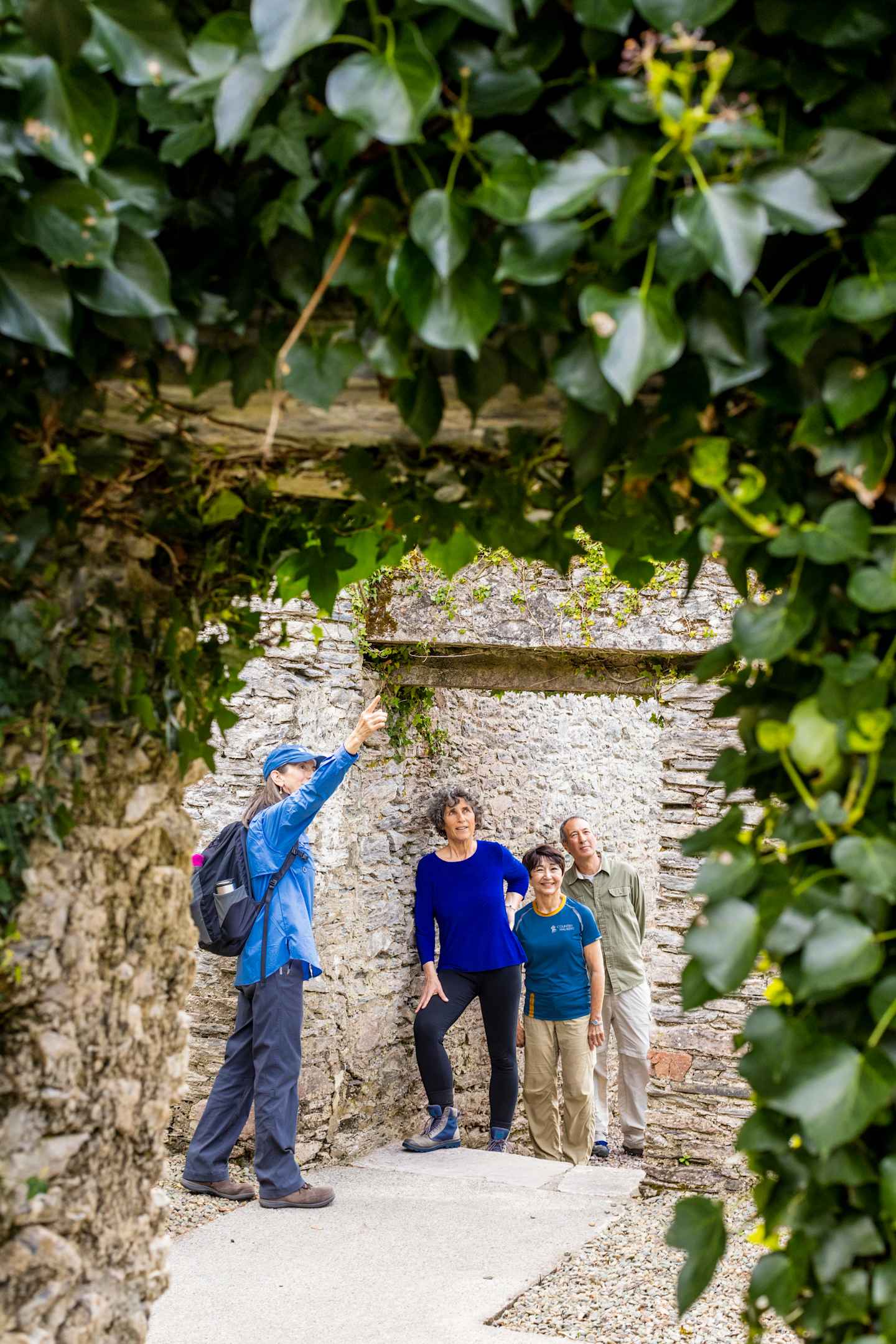 The image shows a group of three people, two adults and one child, walking on a path surrounded by lush greenery and a stone wall in the background.