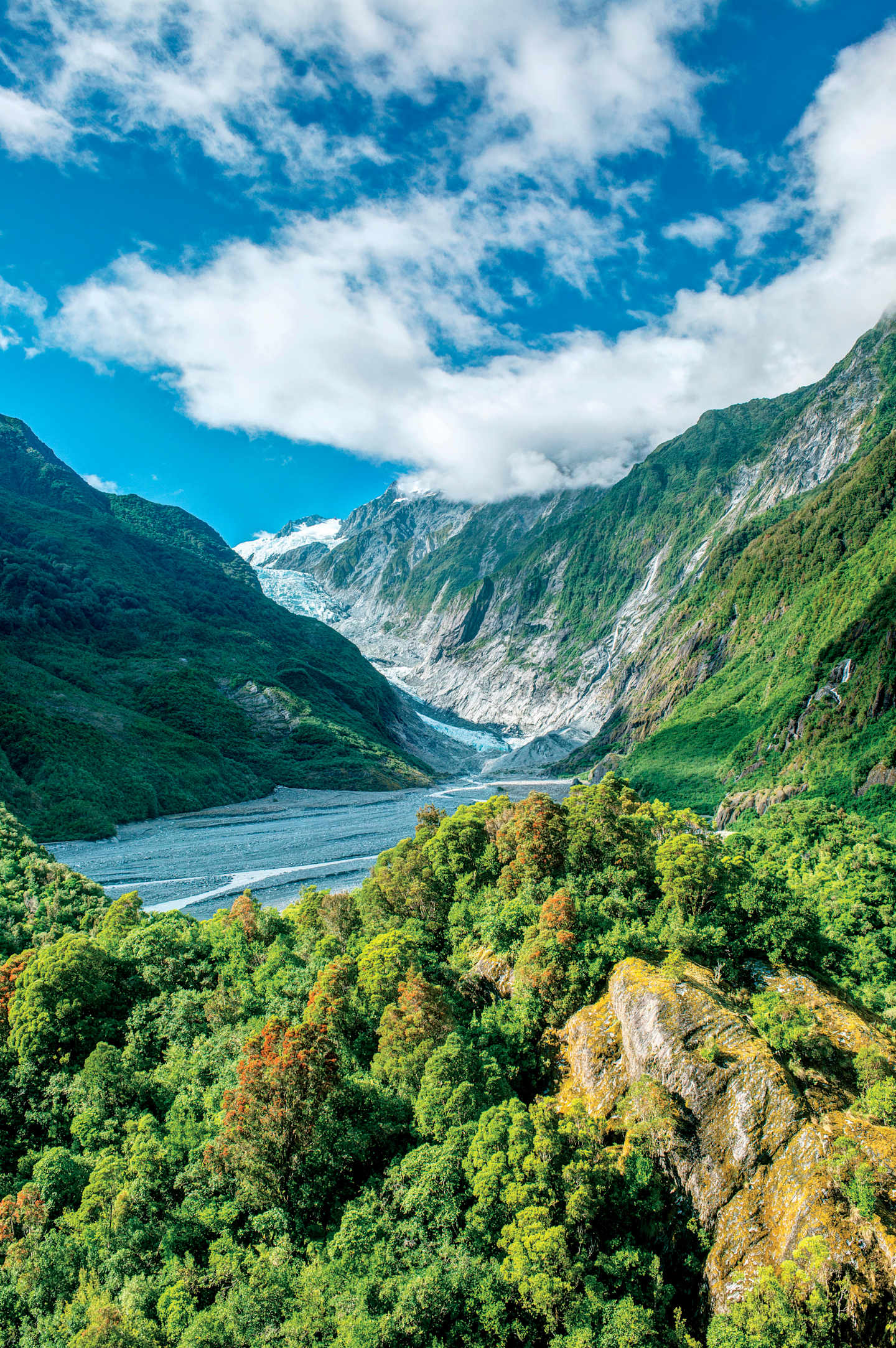 A lush, verdant landscape with a winding river nestled between towering, rugged mountains under a vast, cloudy blue sky.